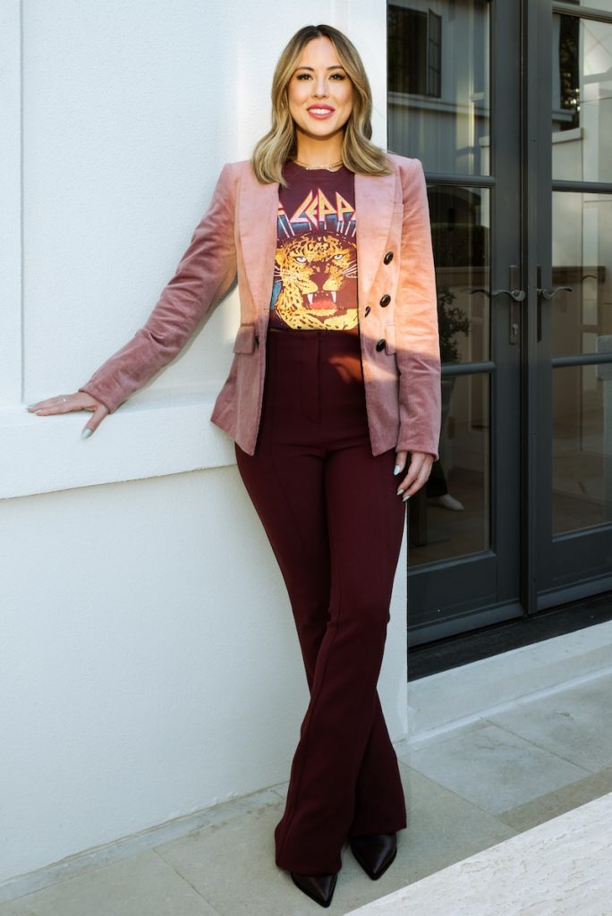 a woman leaning against a wall with her hands on her hips wearing a patterned shirt under a blazer giving off business professional vibes